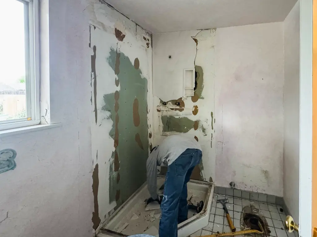 Contractor removing a shower base in a partially demolished bathroom with exposed walls and plumbing