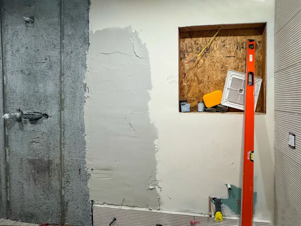 Partially plastered bathroom wall under renovation with exposed wiring, built-in niche, and orange level tool leaning against it.