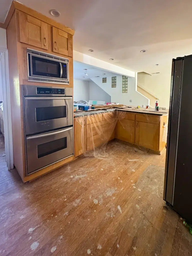 Kitchen under renovation with wooden cabinets, double ovens, and countertops covered in protective plastic.