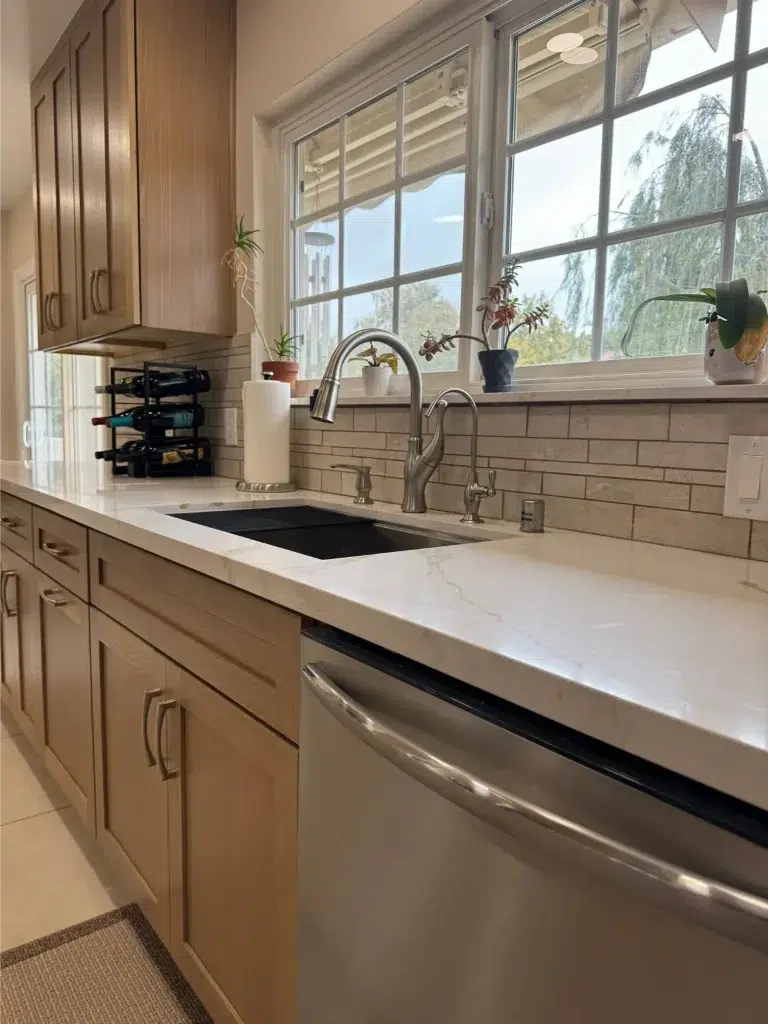 Kitchen sink with stainless steel faucet, light wood cabinets, marble countertop, and potted plants by a window.