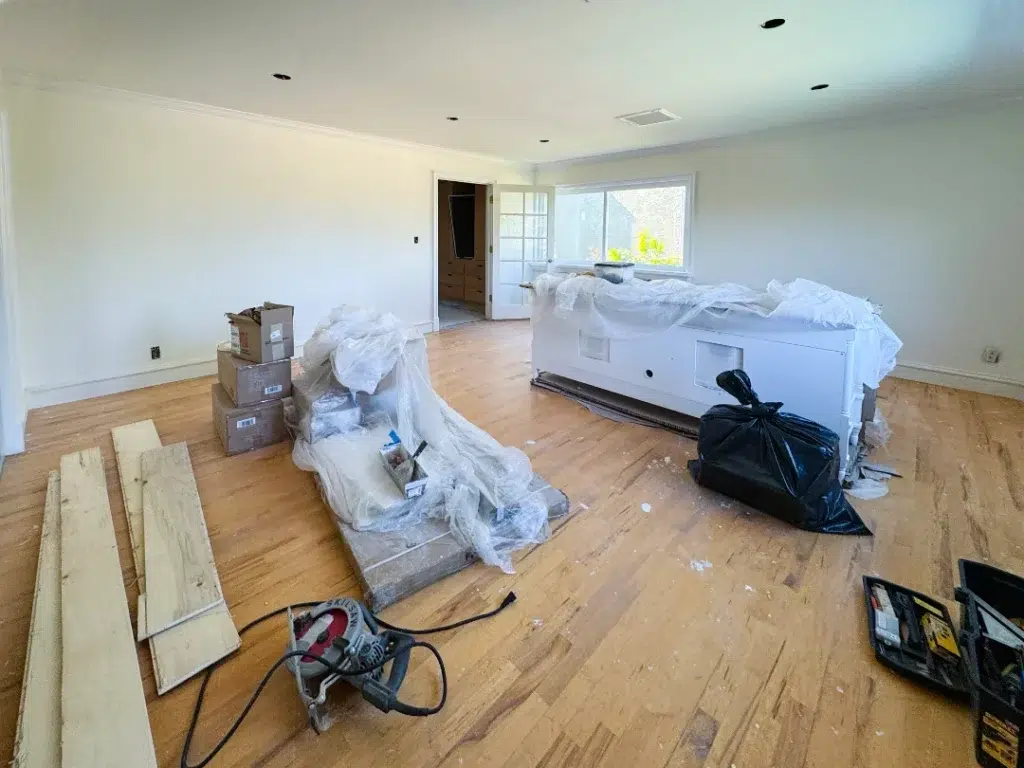 Living room under renovation with flooring materials, tools, and covered furniture spread across the unfinished wood floor.