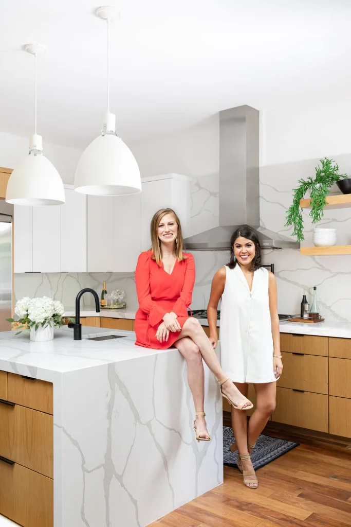 Two women in a modern kitchen with marble waterfall island, wood cabinets, and white pendant lights.