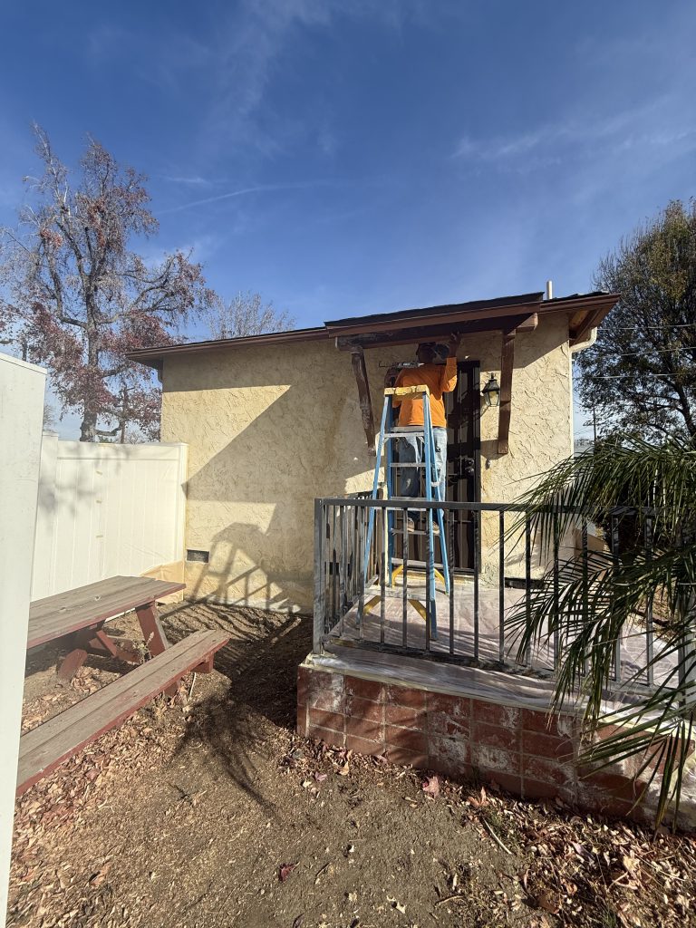 Contractor standing on a ladder repairing a small porch roof overhang on a stucco house exterior