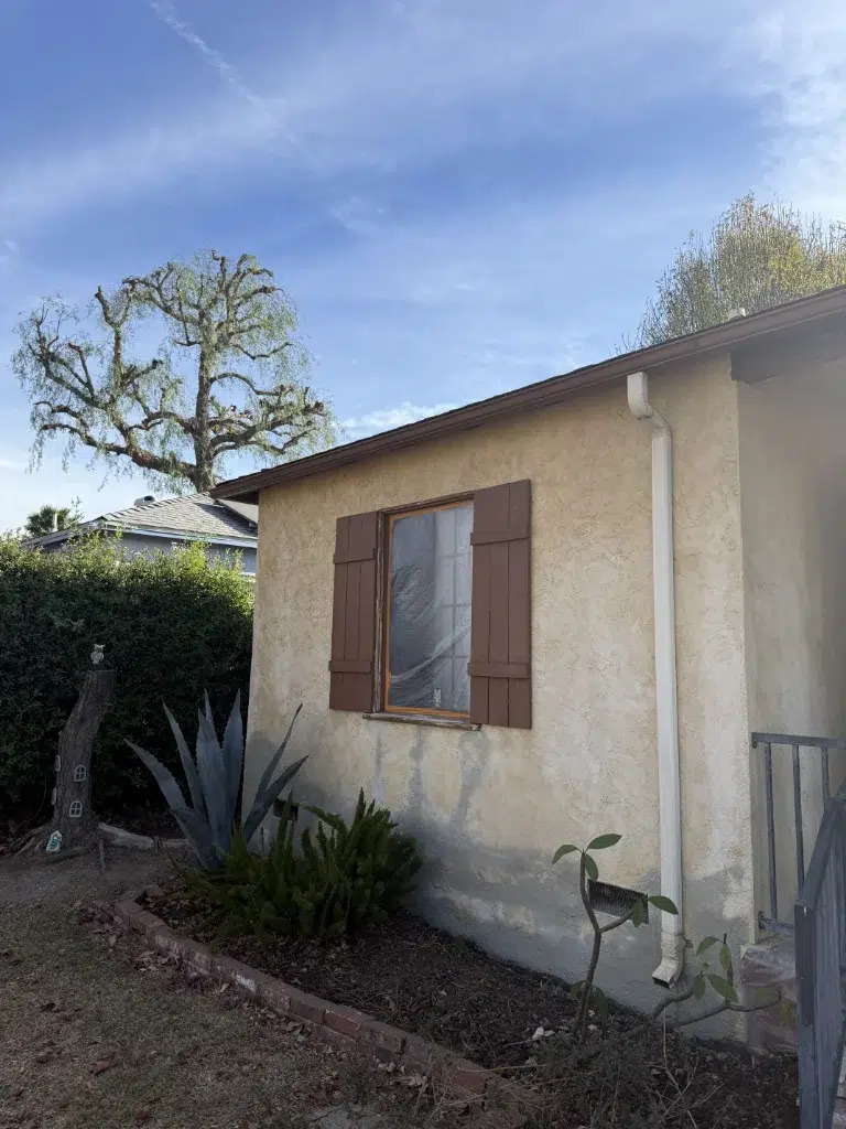 Stucco house wall with brown shutters and landscaping plants beneath a clear sky.