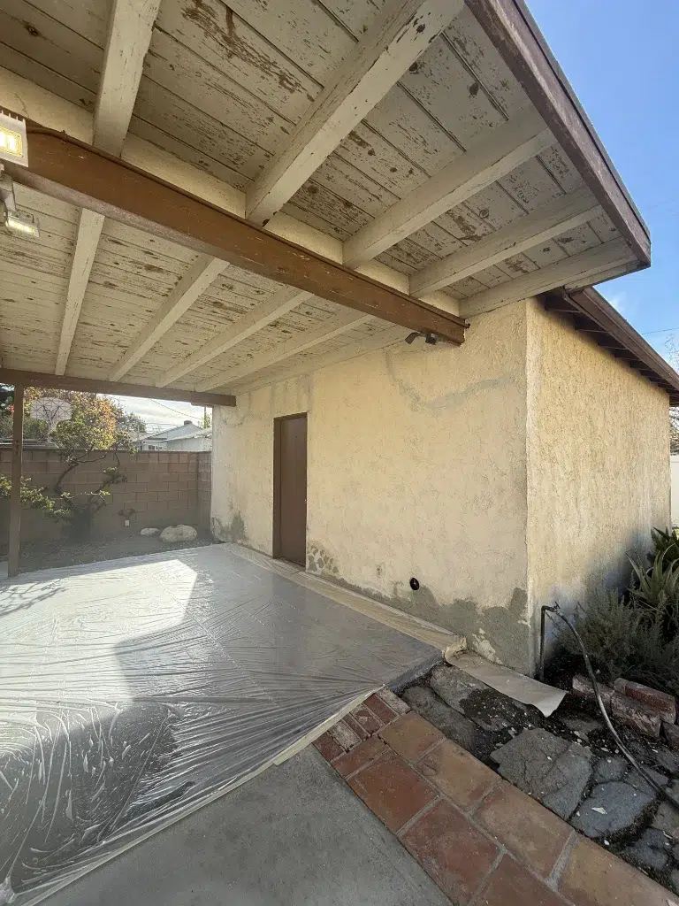 A weathered patio cover with peeling white paint attached to a stucco house, with plastic sheeting on the concrete floor.