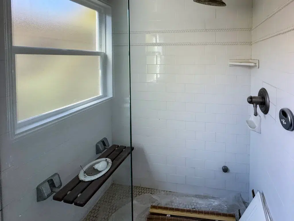White tile shower with a frosted window, wall-mounted bench, hexagon floor tile, and brushed metal fixtures.